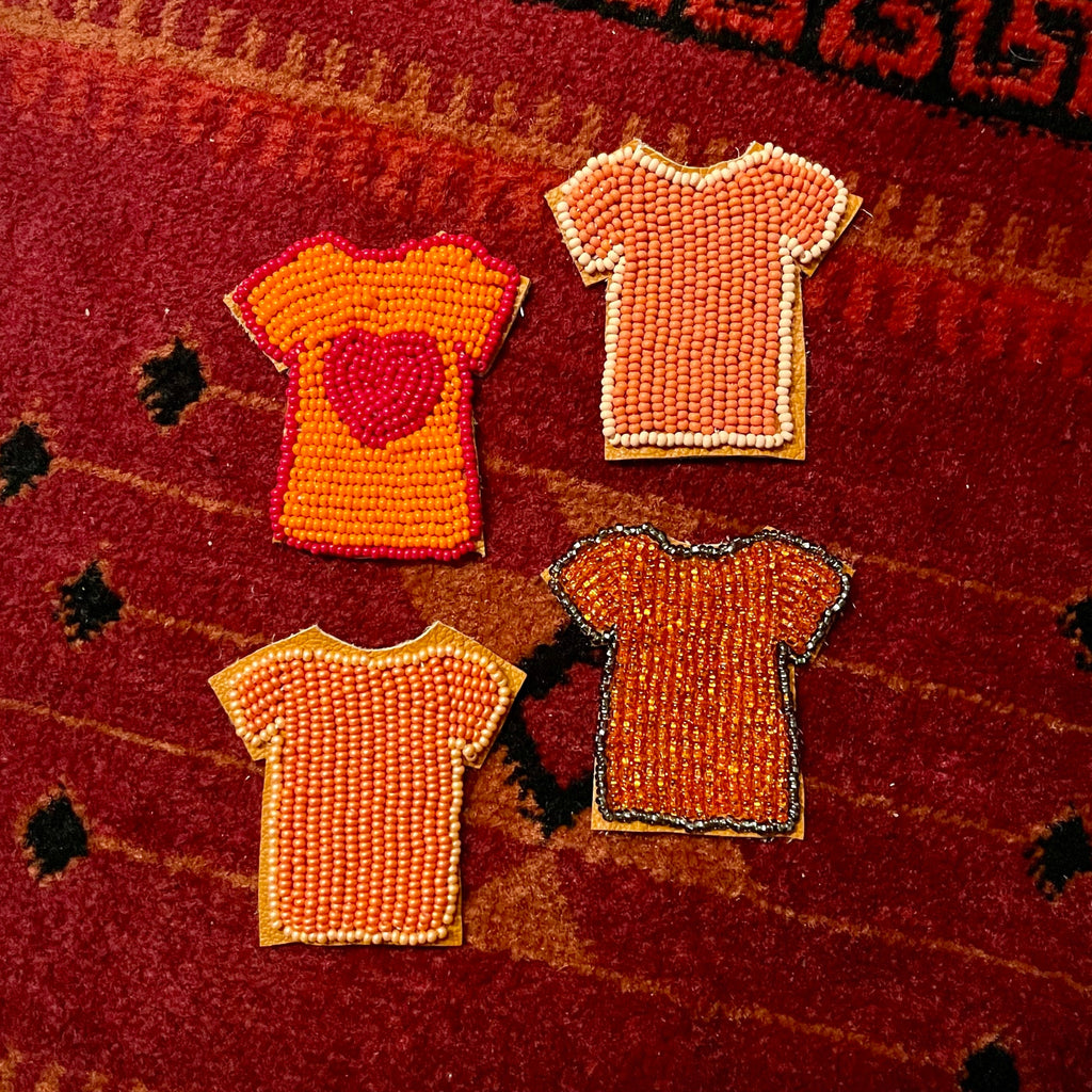 Four small beaded t-shirt appliqués in orange, red, and peach tones arranged on a red patterned textile background.