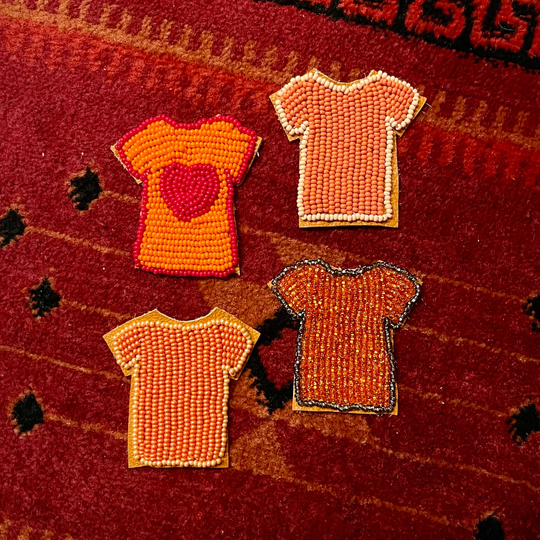 Four small beaded t-shirt appliqués in orange, red, and peach tones arranged on a red patterned textile background.